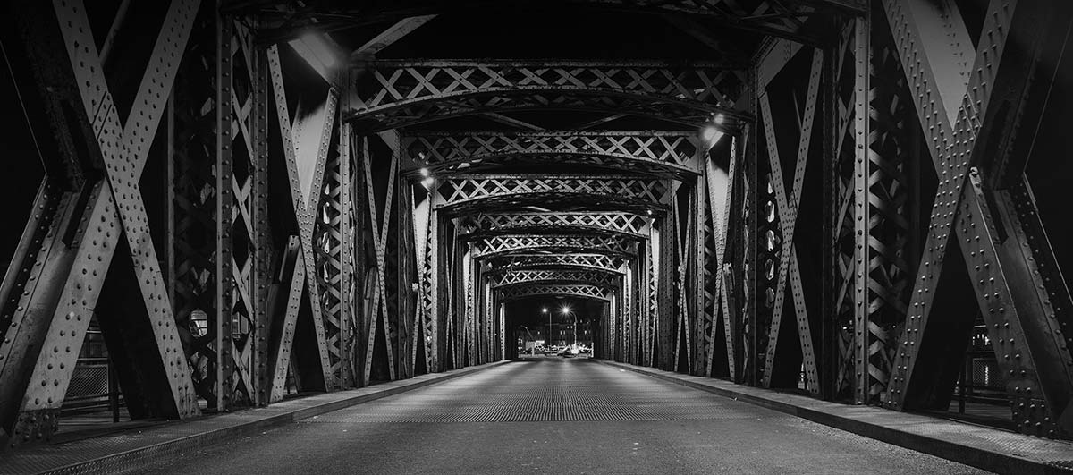 Black and white nighttime photo of a steel truss bridge interior with crisscross beam patterns leading toward distant light